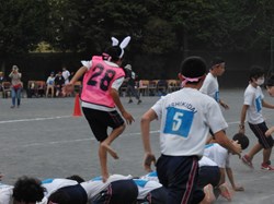 丸まって座った生徒の背中をウサ耳つけた生徒が渡っています