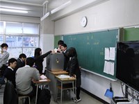 教室で、教卓におかれた投票箱に投票する生徒の写真