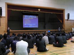 体育館でパワーポイントの画面を見ながら話を聞く生徒